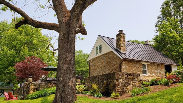 Una casa de piedra con chimenea, rodeada de césped cuidado y árboles.
