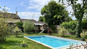 A rectangular swimming pool with a tiled deck surrounding it, a grassy area with various plants, and a wooden play structure in the background. - Charmante Maison Familiale (Gradignan)