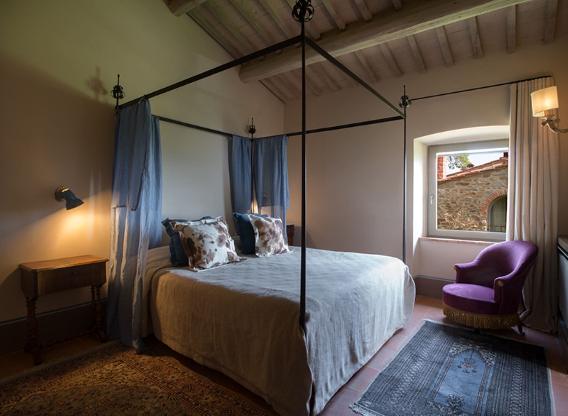 A four-poster bed with a canopy, a wooden nightstand, a purple armchair, and a view of a stone building through a window.