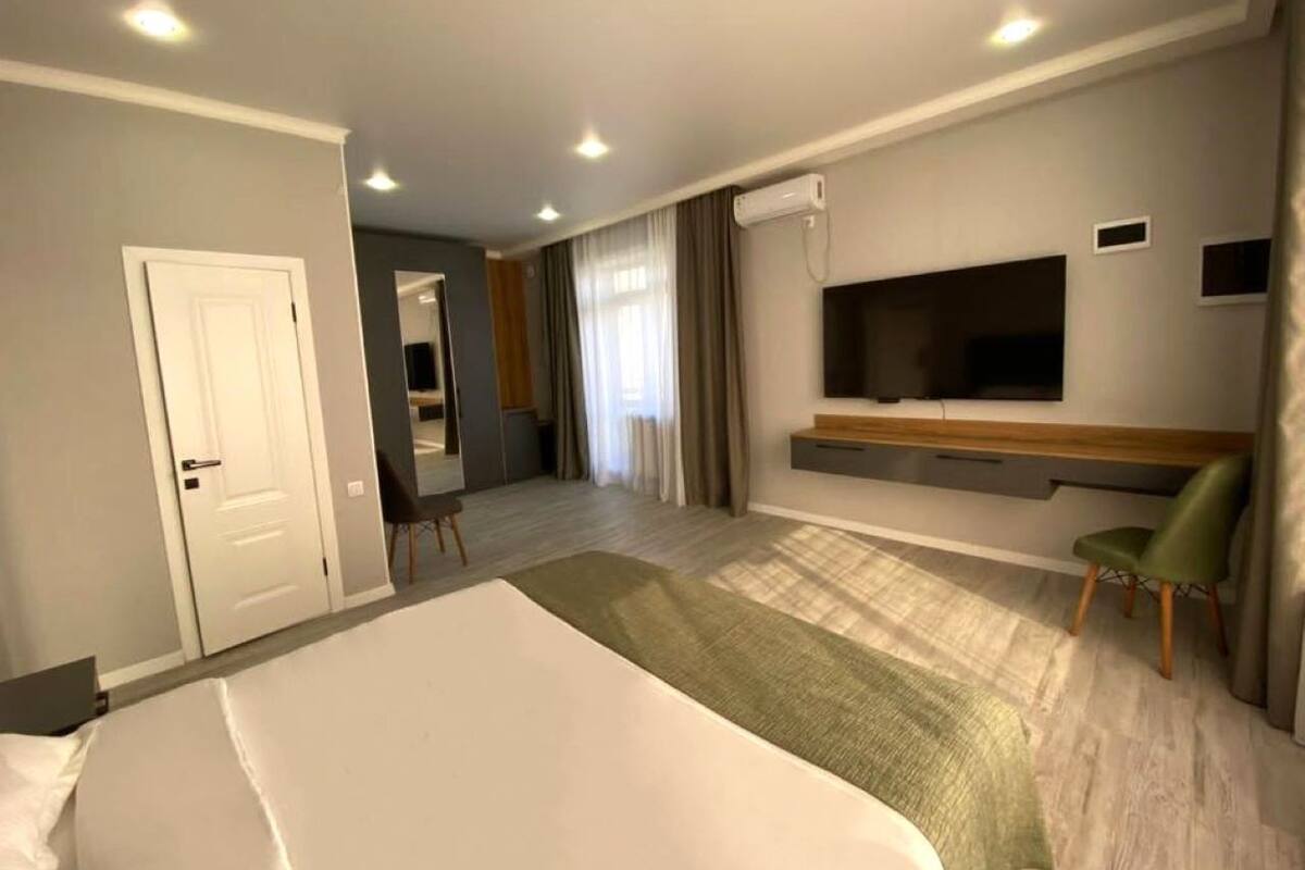 A modern hotel room with a large bed, a flat-screen TV mounted on the wall, a seating area with a chair, and a small desk.