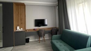 Una habitación de hotel moderna con un sofá verde, un televisor de pantalla plana montado en la pared, un escritorio de madera y un ventanal con cortinas.
