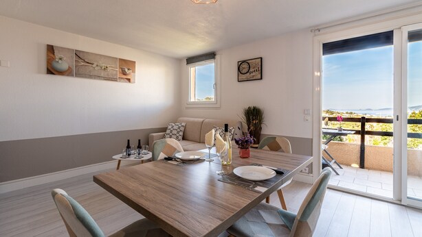 Un coin repas avec une table en bois, des chaises et une vue sur un balcon extérieur, accessible par une porte-fenêtre coulissante.