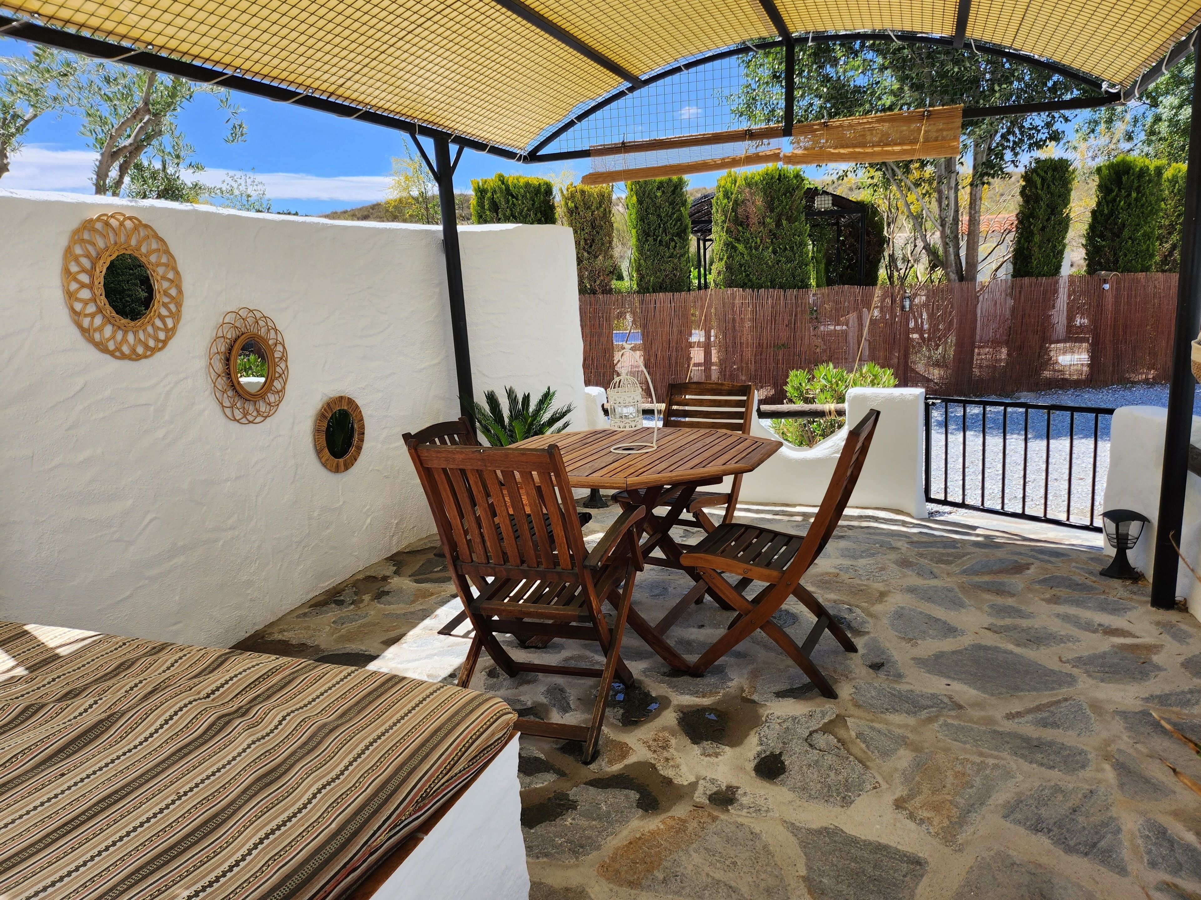 Un patio con una mesa y sillas de madera redonda, un área cubierta con un toldo amarillo y un suelo de piedra.