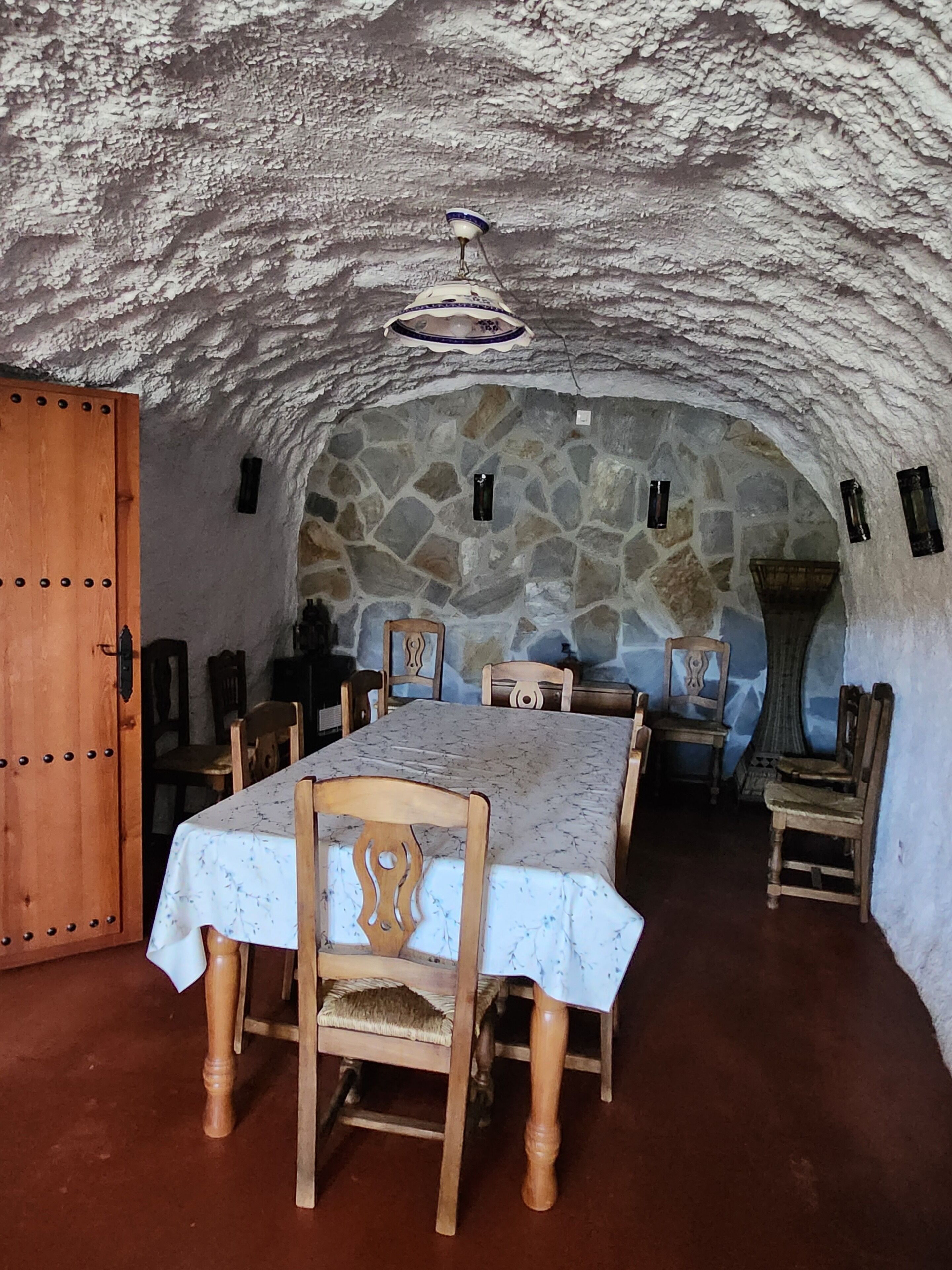 A dining area with a stone wall, wooden chairs, a table with a floral tablecloth, and a hanging light fixture.