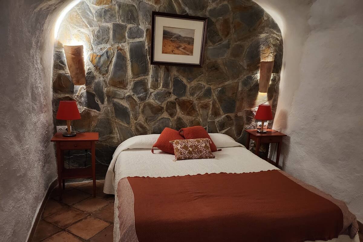 Un dormitorio acogedor con pared de piedra, una cama con almohadones rojos y estampados, y dos mesitas de noche con lámparas.