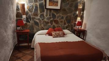 Une chambre confortable avec un mur en pierre, un lit agrémenté d’oreillers rouges et à motifs, et deux tables de chevet avec des lampes.