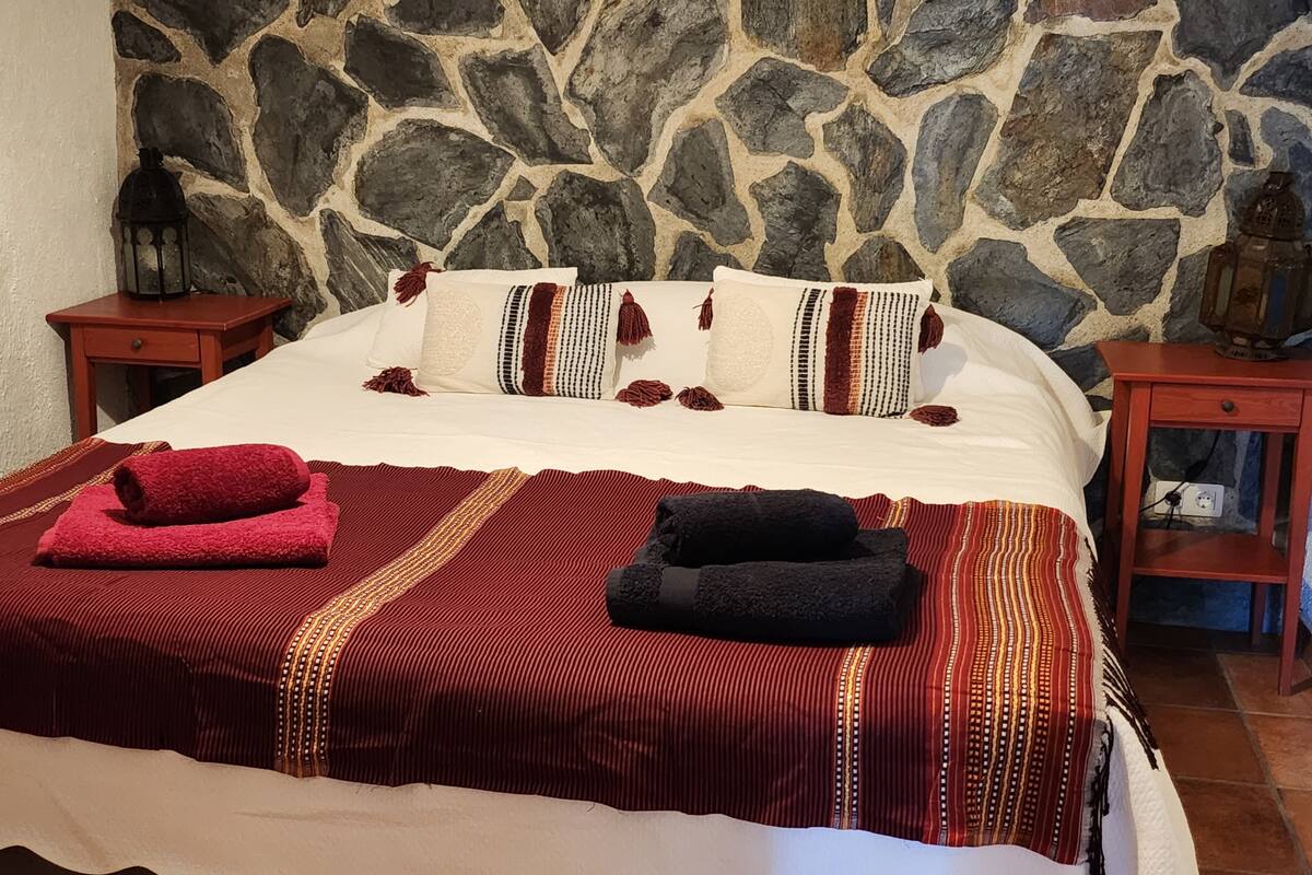 Una cama con un fondo de pared de piedra, dos mesitas de noche con faroles y una colcha a rayas dorada y roja.