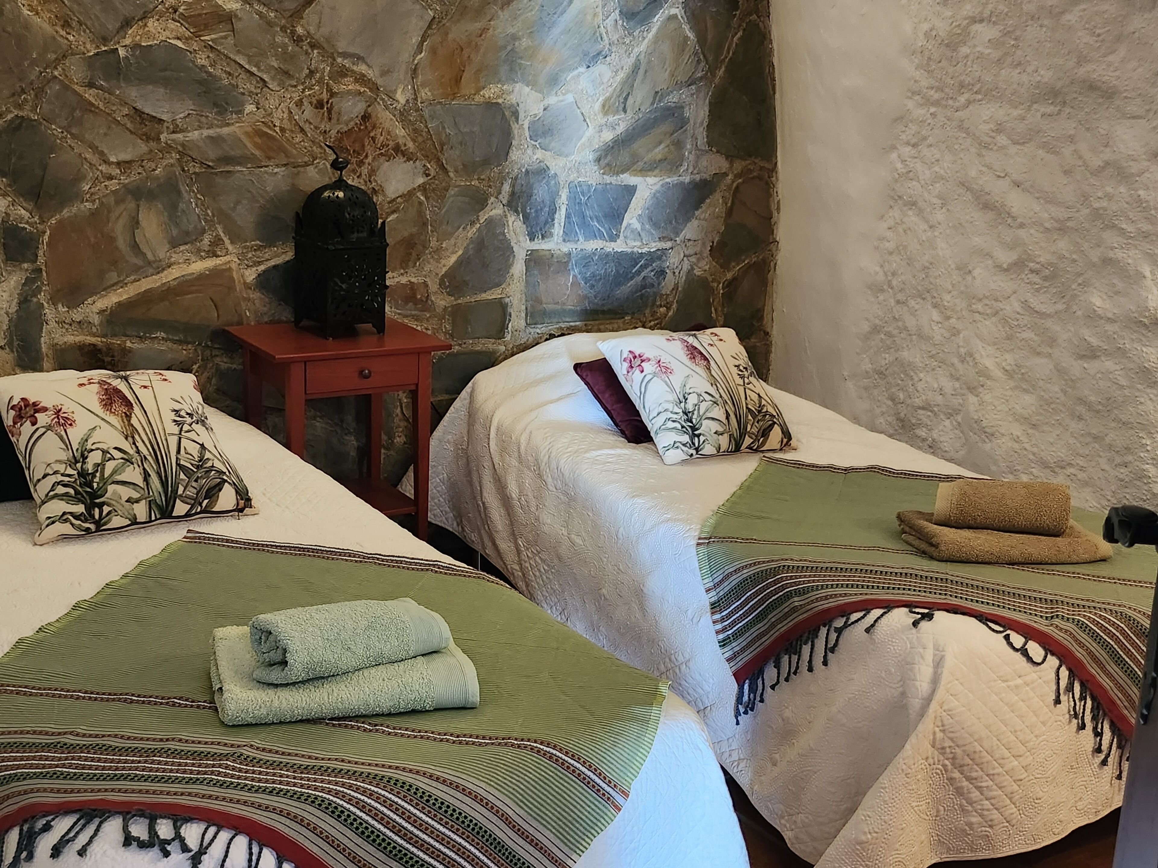 Una habitación con dos camas, cada una con una manta verde y blanca, una mesita con un florero y una pared de piedra.