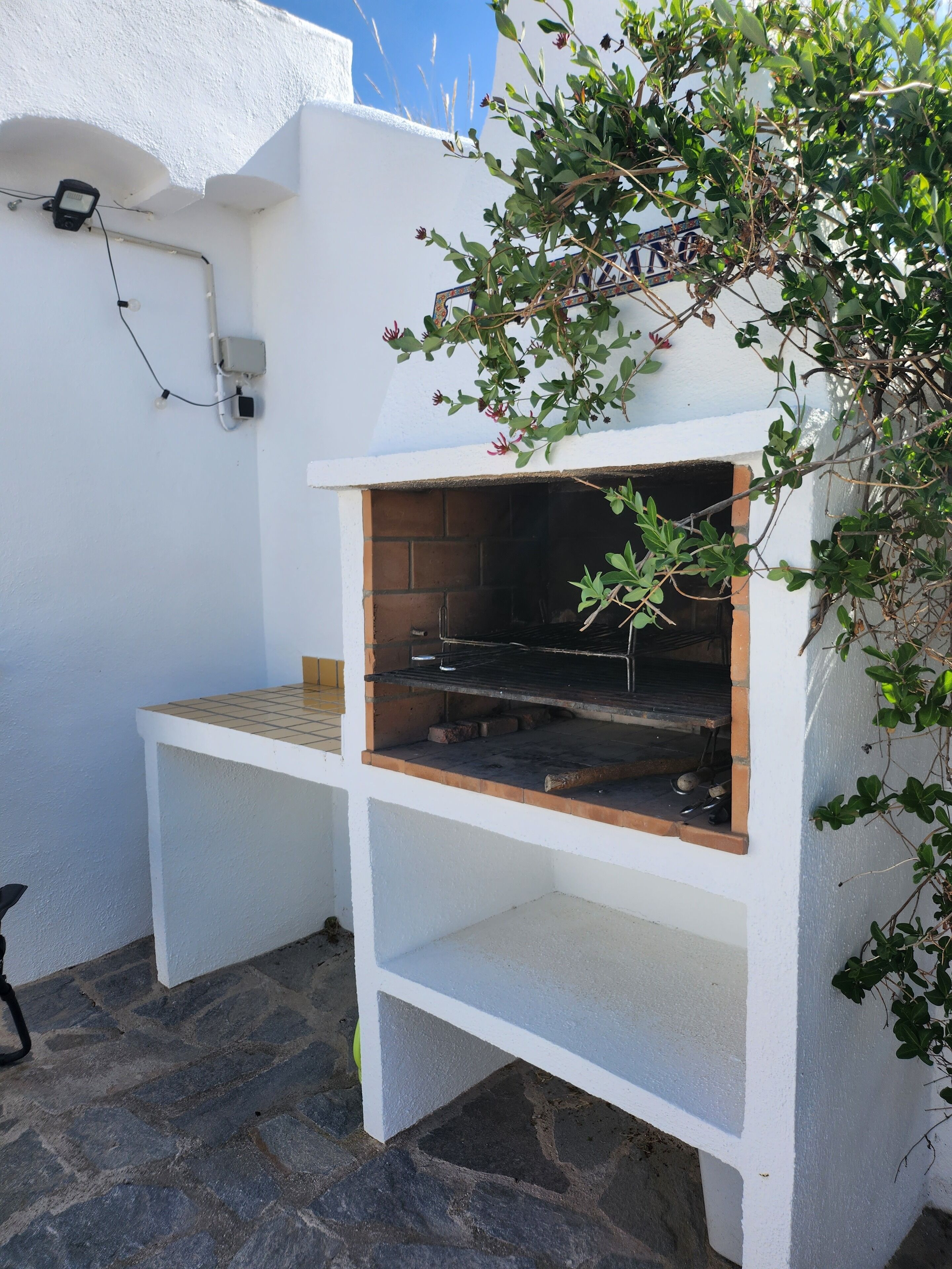 A white outdoor grill with a tiled top and a brick base, set against a white wall with a small window and a power outlet.