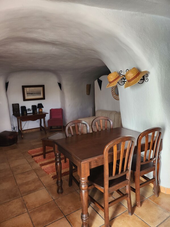 Un área de comedor con una mesa y sillas de madera, una silla roja, una mesita con una radio y una lámpara, y un cuadro en la pared.