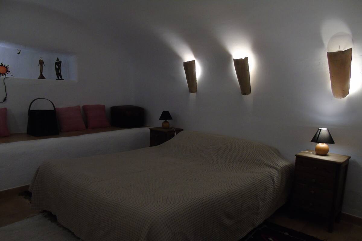 Un dormitorio tenuemente iluminado con una cama grande, una mesita de noche con lámpara y apliques en la pared.