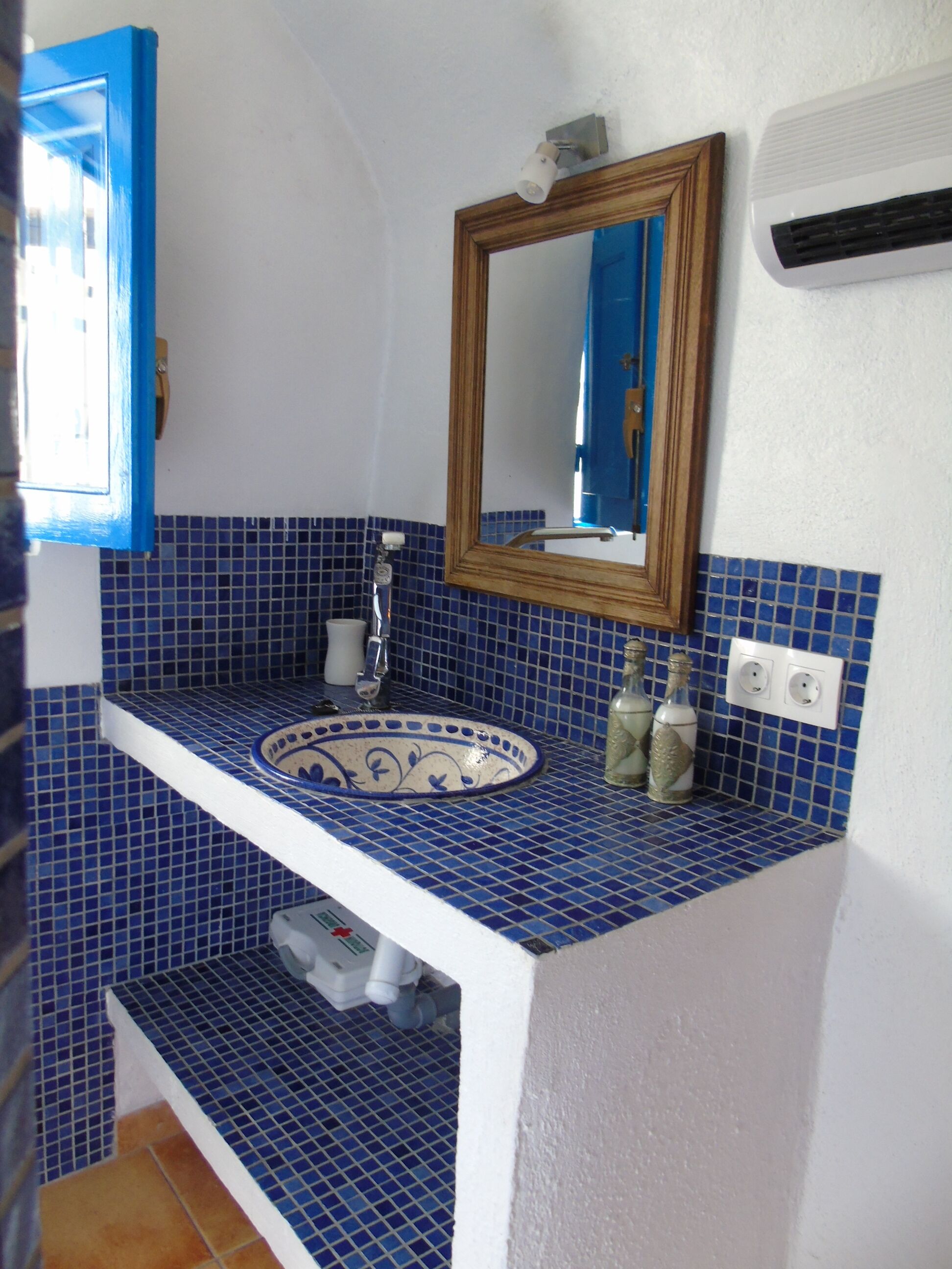 Een badkamer met een blauwe betegelde spatwand, een wit wastafelmeubel met decoratieve kom, een spiegel met houten lijst, en een witte airconditioningunit aan de muur.