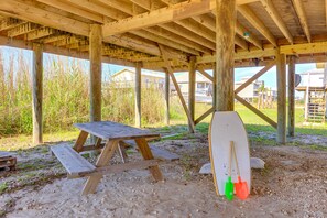 A wooden picnic table under a shelter with a surfboard and shovels beside it. - Breezy Dauphin Island Vacation Rental with Deck! (Dauphin Island)