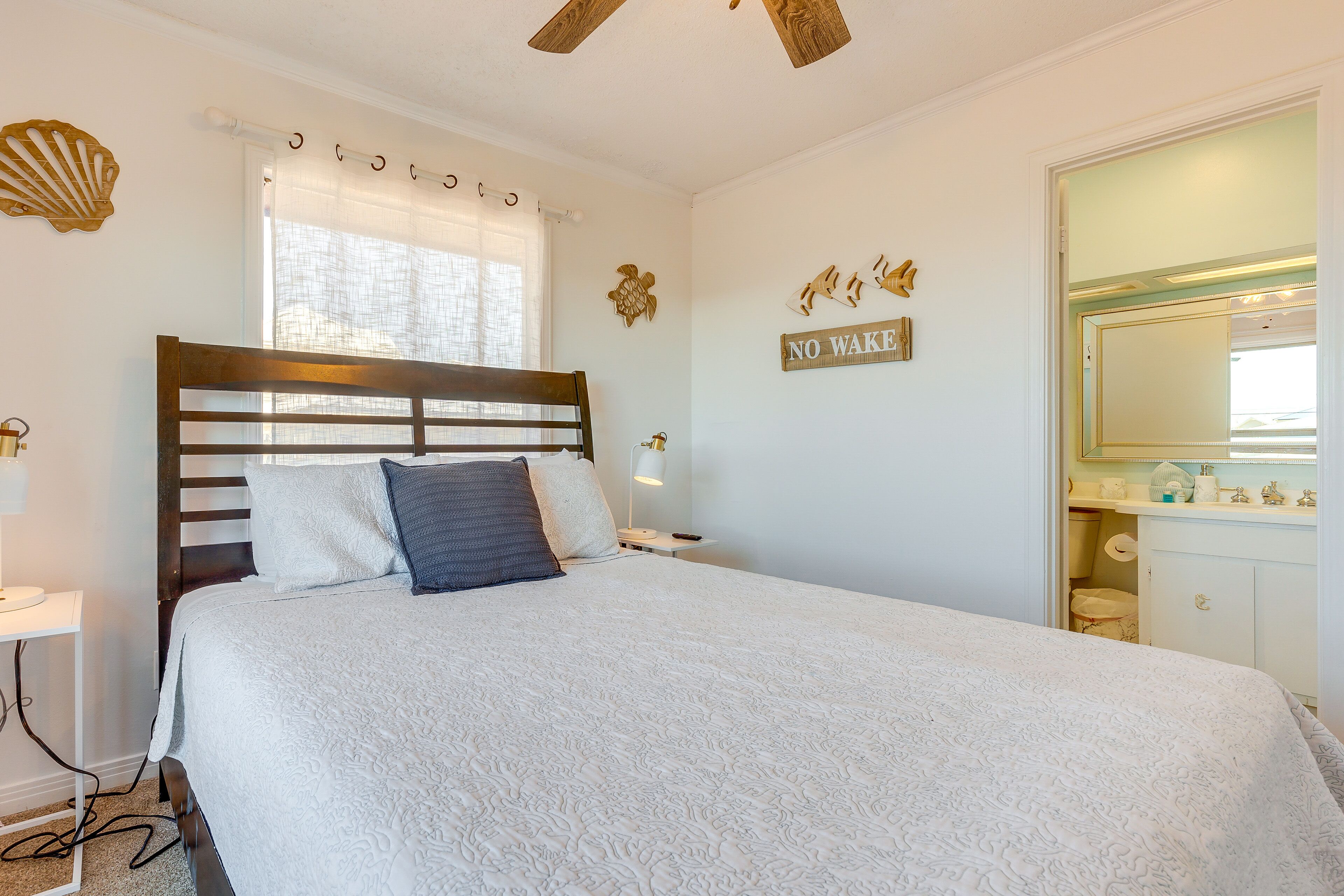 Un dormitorio con cabecera de madera, cama con ropa de cama blanca, mesita de noche, lámpara, ventana con cortinas, un reloj de pared, y un baño visible a través de una puerta abierta.