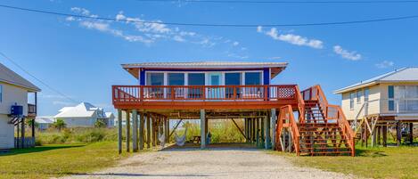 Una casa frente a la playa con un balcón rojo y escaleras de madera que conducen a una terraza cubierta.