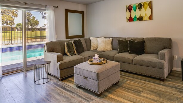 A living room with a sectional sofa, a small ottoman, and a side table. There is a large window with a view of a swimming pool and a fence. - Pool & Spa: Palm Desert Home Near Golf! (Palm Desert)