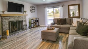 A living room with a stone fireplace, a flat-screen TV, a ceiling fan, and a large window with a view of the outdoors. - Pool & Spa: Palm Desert Home Near Golf! (Palm Desert)