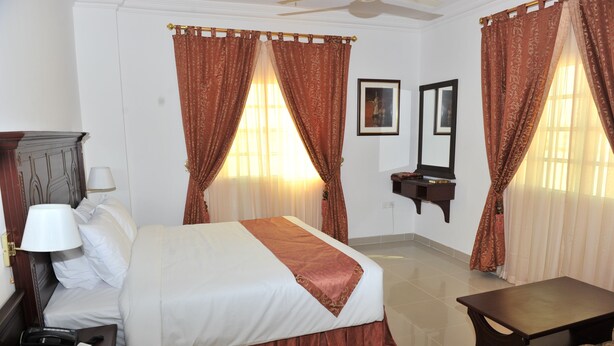 A hotel room with a bed, a nightstand with a lamp, a bedside table, a small table, and a mirror on the wall. - Manam Sohar Hotel Apartments (Sohar)