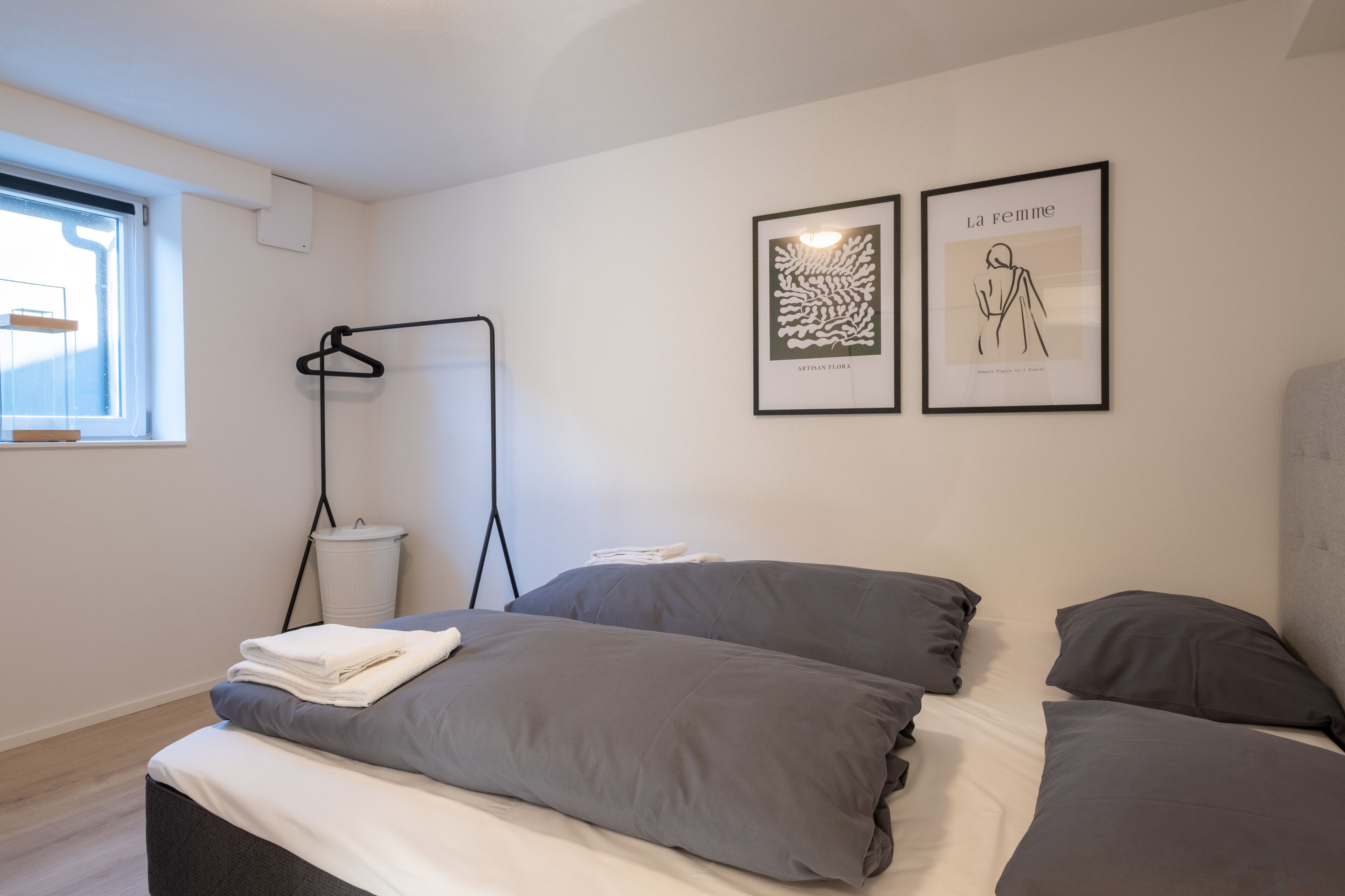 Une chambre moderne avec un lit, une literie grise, une table de chevet blanche, une lampe suspendue et deux œuvres d’art encadrées accrochées au mur.