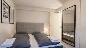 Un lit bien fait, avec une literie grise, une tête de lit grise et un grand miroir au mur.