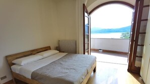 Ein Schlafzimmer mit einem Holzbett, Blick auf einen See durch eine offene Tür und ein Balkon mit Bergblick.