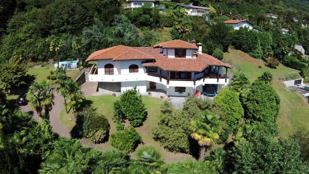 Ein großes, zweistöckiges Haus mit Ziegeldach, umgeben von üppiger Vegetation und weiteren Häusern im Hintergrund.