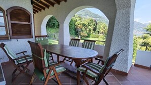 Eine überdachte Terrasse mit einem runden Tisch und sechs Stühlen, mit Blick auf eine üppige Landschaft und Berge.