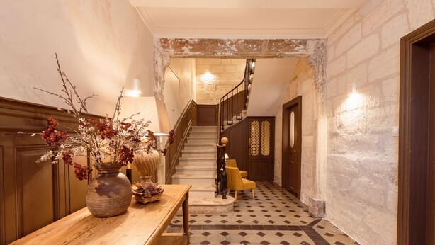Ein Flur mit einer hölzernen Bank, einer Vase mit getrockneten Blumen, einer Treppe und einem gemusterten Boden.