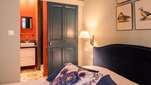Ein Schlafzimmer mit dunkelblauer Kopfteil, einem Bett mit floraler Bettwäsche, einem Nachttisch mit Lampe, einem Kleiderschrank und einem durchgegangenen Türrahmen einsehbaren Badezimmer.