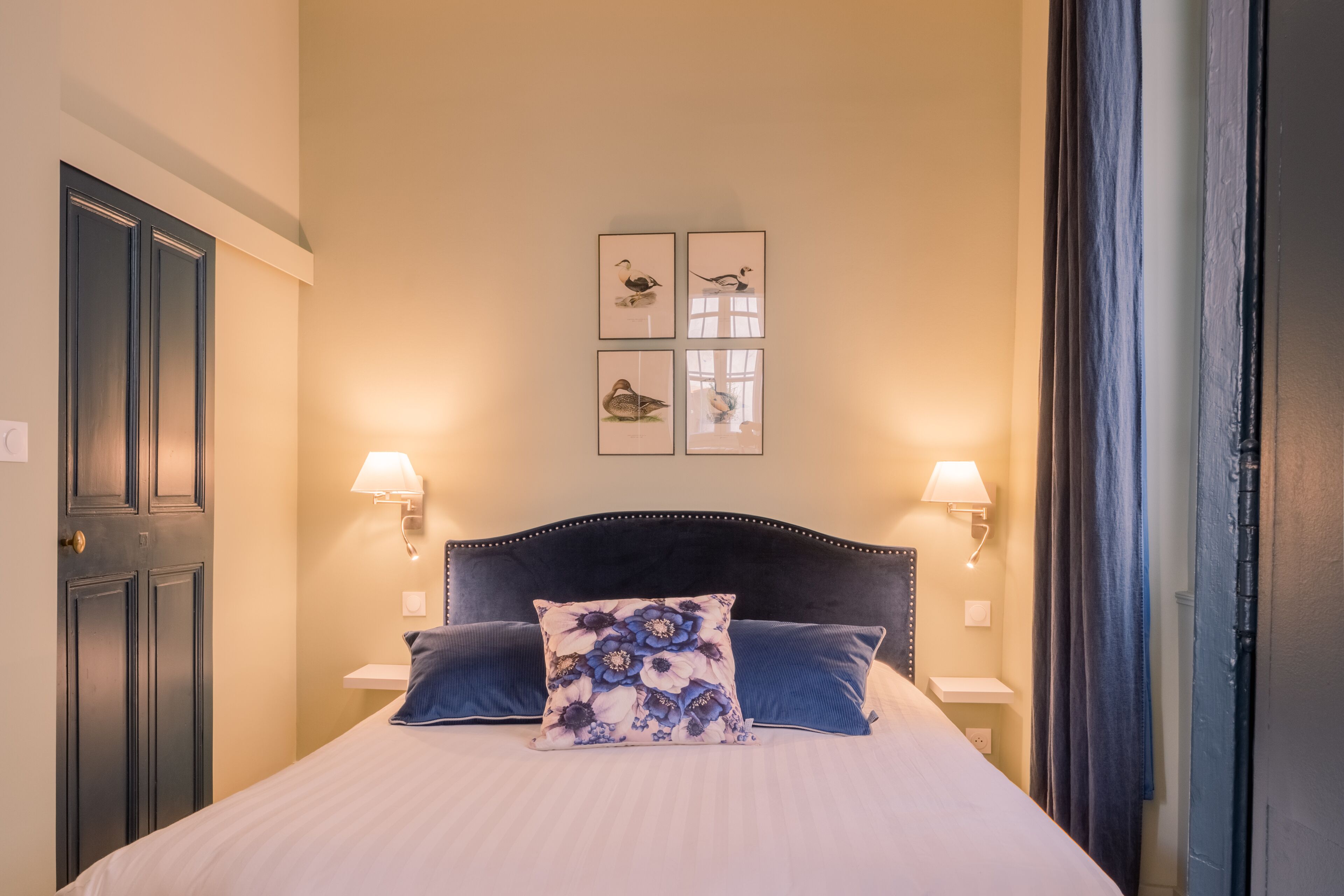 A bedroom with a bed, two wall-mounted lamps, a dark headboard, and framed artwork on the wall.