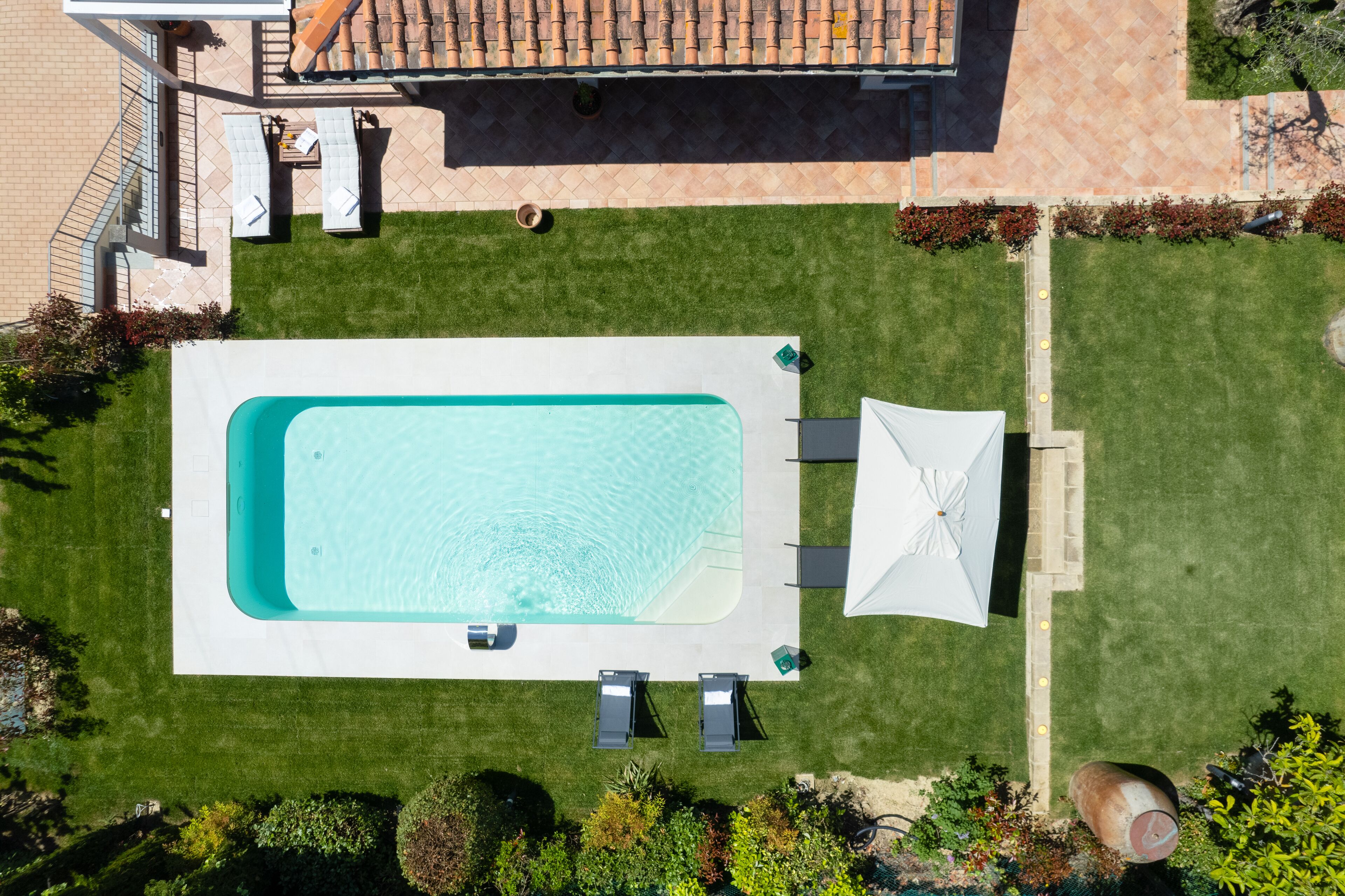 An aerial view of a swimming pool with a white deck, surrounded by a well-maintained lawn and a patio area with outdoor furniture.