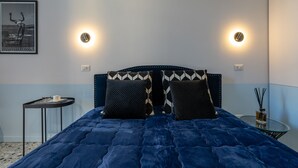 A bedroom with a dark blue quilted bedspread, two patterned pillows, a small round table with a candle, and a wall-mounted picture. - Teatro Toledo 272 (Naples)
