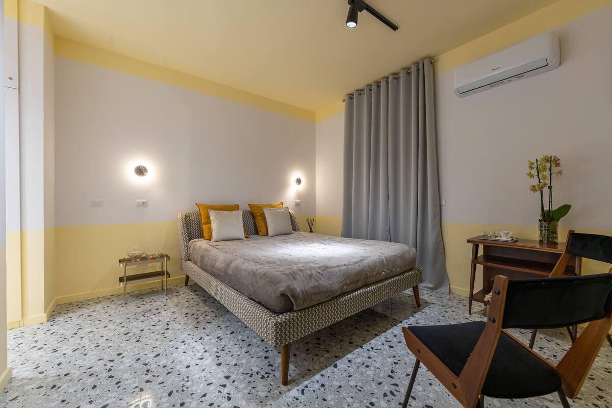 A bedroom with a bed, bedside table, chair, and an air conditioning unit.