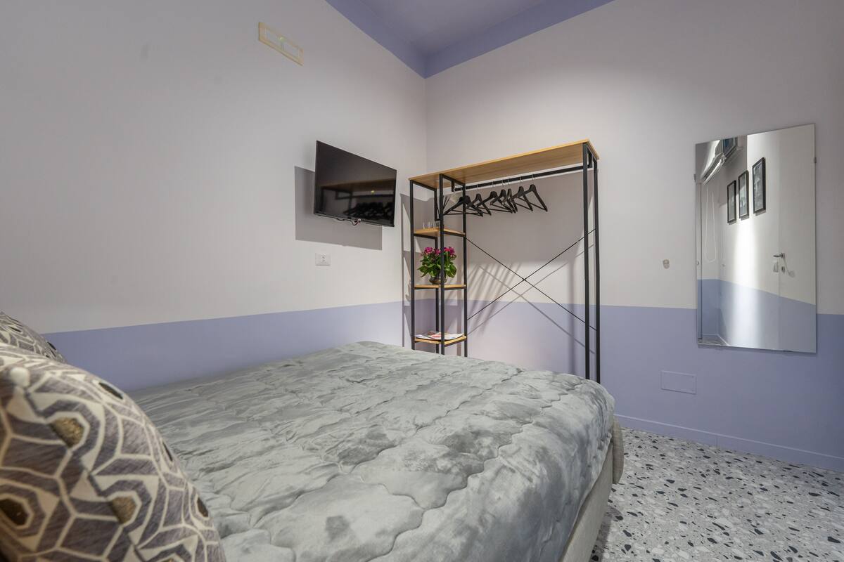 A bedroom with a bed, a TV mounted on the wall, a clothes rack, and a mirror.