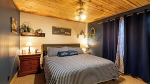 A bedroom with a wooden ceiling, a bed with striped bedding, two bedside tables with lamps, a wall-mounted shelf with decorative items, and a dark curtain.