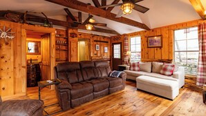 A cozy log cabin interior with a brown leather sofa, a white sofa, wooden floors, and a ceiling fan.