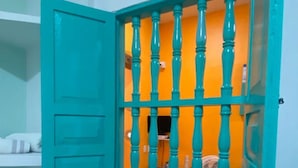 A room with a teal door and window, a bed with a teal pillow, and a small table with a fan.