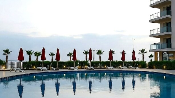 Una piscina con sillas de descanso y sombrillas, rodeada de palmeras y un edificio con balcones.
