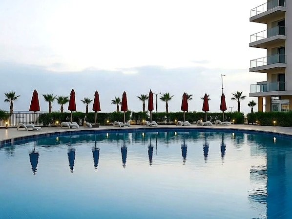 A swimming pool with lounge chairs and umbrellas, surrounded by palm trees and a building with balconies.