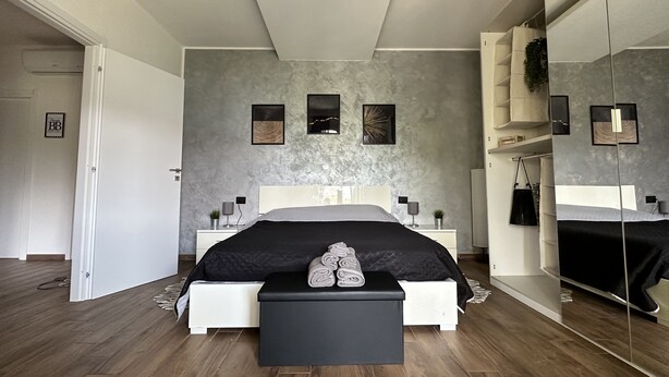 Een moderne slaapkamer met een groot bed, een nachtkastje, een zwarte bank, een grote spiegel en een gestructureerde grijze muur.