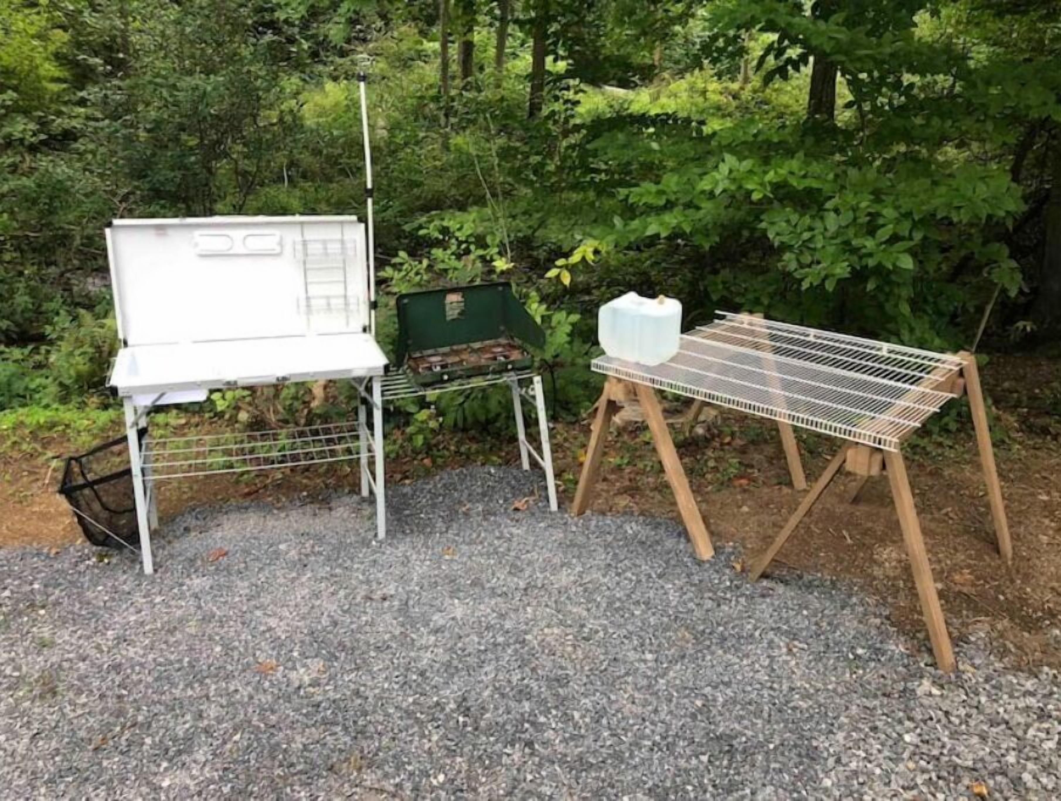 Ein tragbarer Grill mit Metallgestell und weißem Behälter auf einem Gestell, umgeben von Kies und Grünpflanzen.