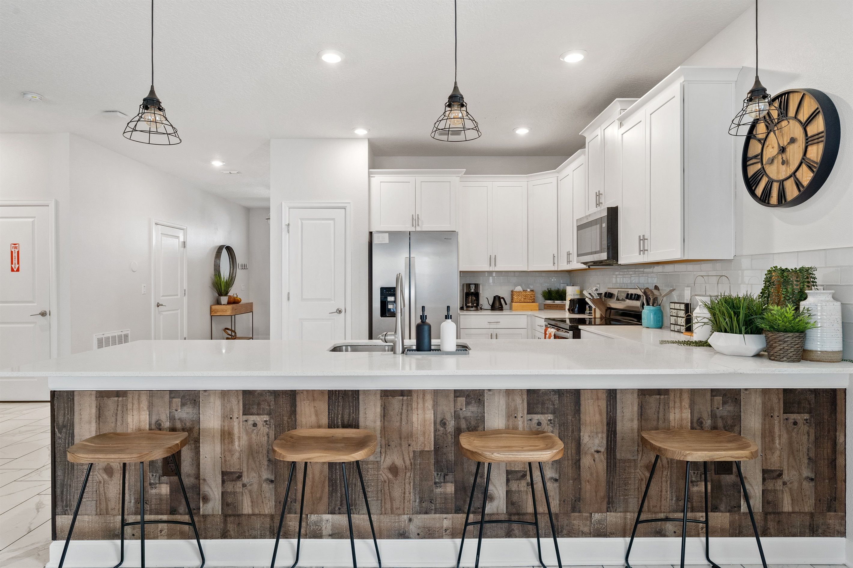 A modern kitchen with white cabinets, a central island with stools, pendant lighting, and a wooden backsplash.