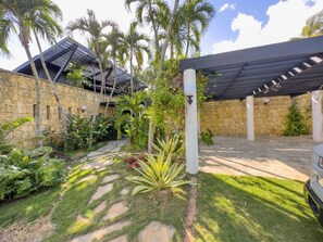 Um edifício de paredes de pedra com um alpendre coberto, rodeado por vegetação exuberante e palmeiras.
