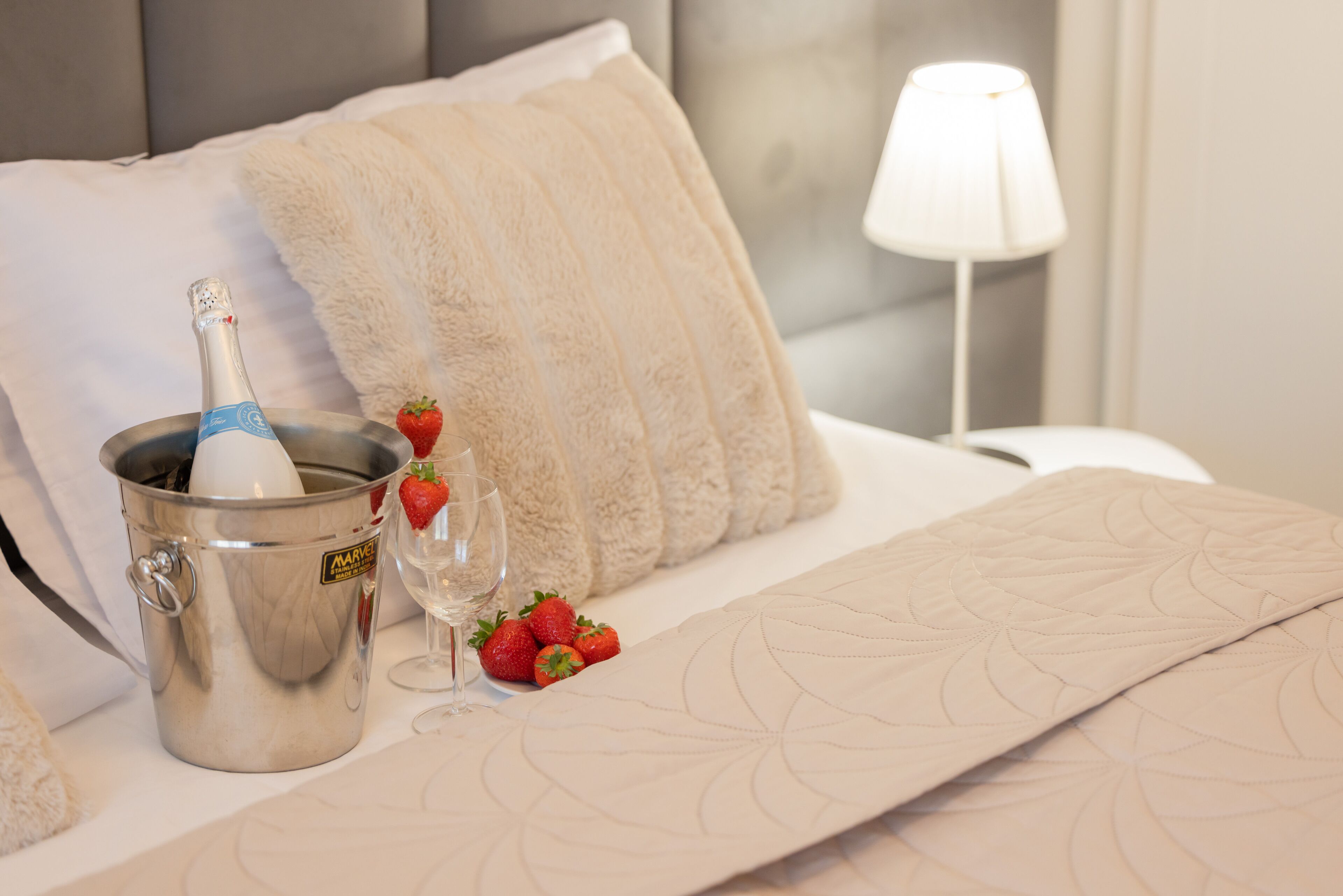 A bed with a white quilt, a champagne bucket, a glass, and strawberries.