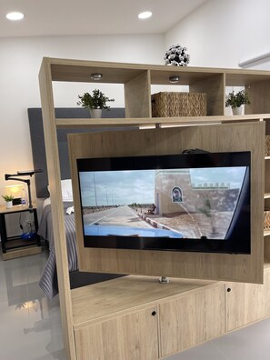 Ein modernes Wohnzimmer mit einem großen Flachbildfernseher, der an einer Holz-Unterhaltungseinheit befestigt ist. Auf der Einheit stehen zwei Zimmerpflanzen und auf einem Beistelltisch steht eine Lampe.
