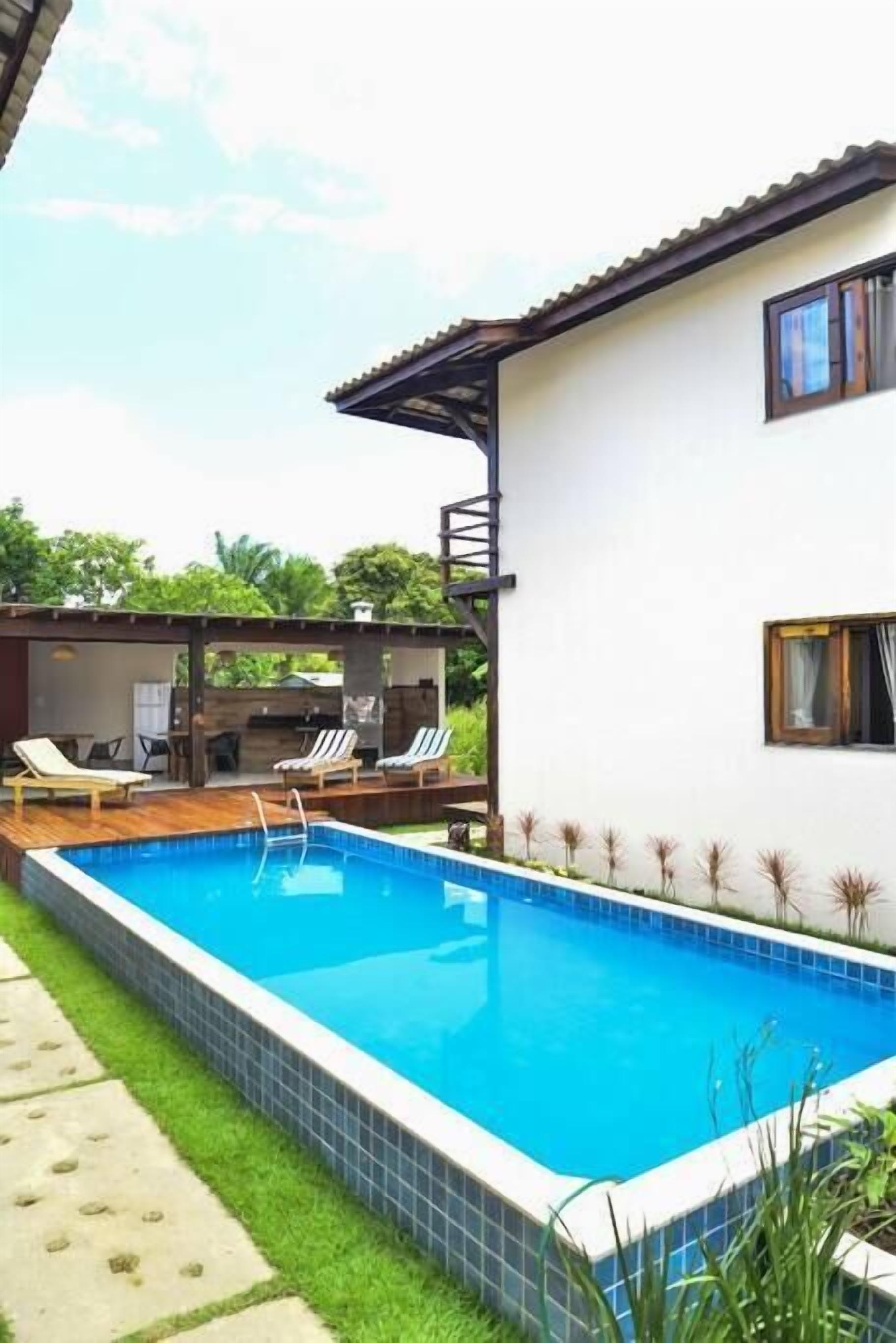 A swimming pool with a wooden deck and lounge chairs, surrounded by a white building with a tiled pool edge.