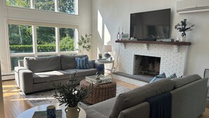 Un soggiorno moderno con un divano grigio, un caminetto bianco, una TV a schermo piatto e ampie finestre che si affacciano sul verde.