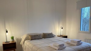 Un lit bien fait avec deux oreillers, une table de chevet avec une lampe et une fenêtre donnant sur des arbres.