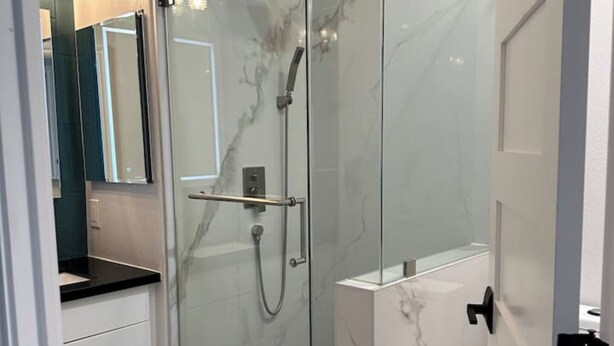 A modern bathroom with a glass-enclosed shower, marble walls, a black countertop, and a teal rug.