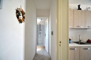 A narrow hallway leading to a bathroom with a shower, a kitchen area with cabinets and a sink, and a decorative wreath on the wall. - Alba Two-room Air-conditioned Holiday Home for 4 Guests (Melendugno)